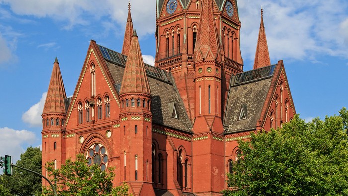 Große rote Kirche mit mehreren Türmen und spitzen Bögen, umgeben von Bäumen und einem klaren blauen Himmel.