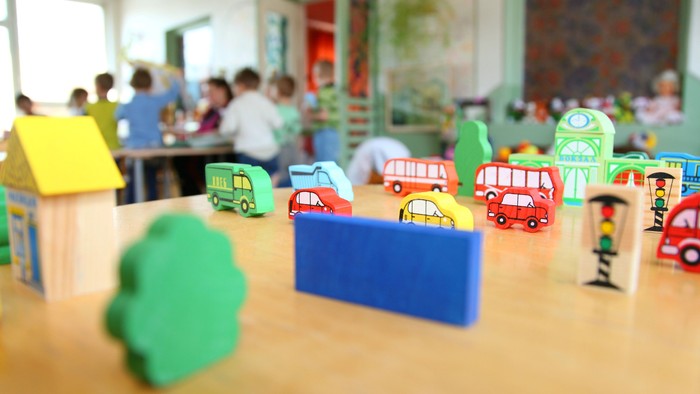Im Vordergrund des Bildes stehen kleine Holzspielzeuge, zum Beispiel Autos und Häuser, auf einem Tisch. Im Hintergrund sieht man Kinder um einen Tisch stehen und sitzen.