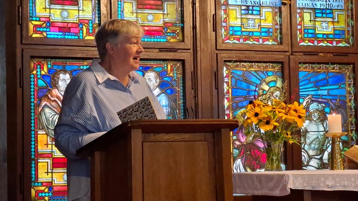 Eine Frau mit Buch an einem Pult vor bunten Mosaikfenstern.