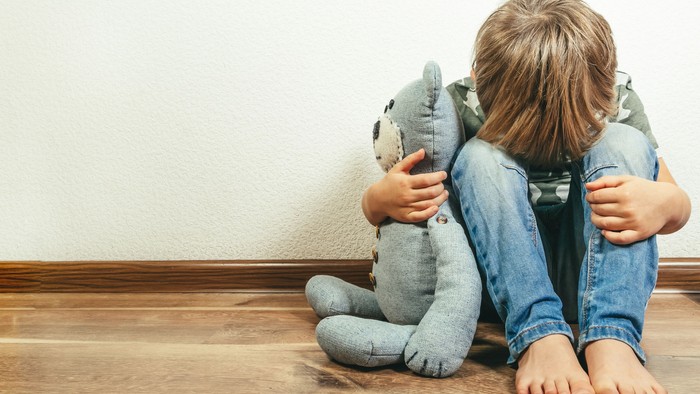 Ein kleiner Jungs sitzt mit gesenktem Kopf auf dem Boden. Sein Gesicht ist nicht zu sehen. Seine Stirn liegt auf seinen Knien. Mit seinem rechten Arm umfasst er ein Kuscheltier. Die linke Hand liegt auf seinem Bein.