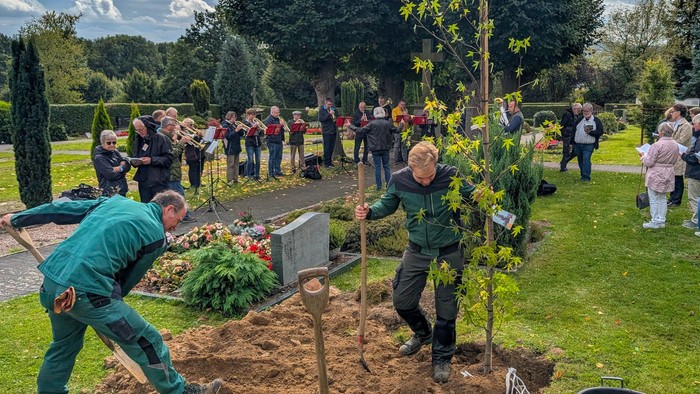 Zwei Gärtner pflanzen einen Spitzahorn auf einem Friedhof. Im Hintergrund spielt ein Posaunenchor und Menschen singen.
