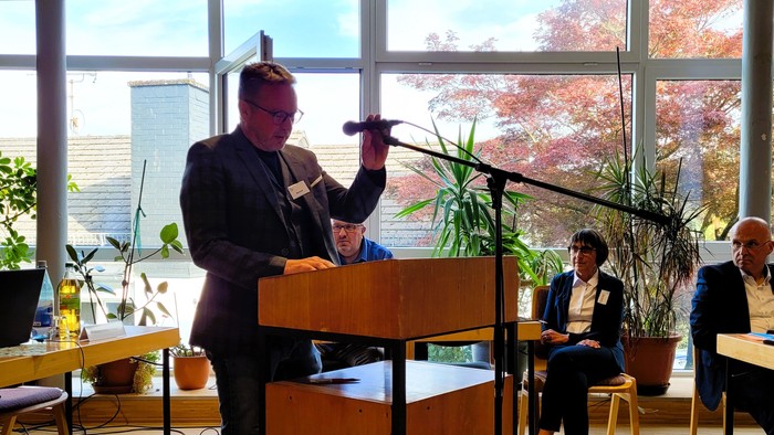 Ein Mann steht an einem Rednerpult und richtet das Mikrofon, hinter ihm große Glasfenster mit Grünpflanzen und Menschen auf Stühlen, die ihm aufmerksam zuhören.