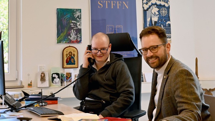 Zwei Männer sitzen an einem Schreibtisch, einer telefoniert, der andere schaut zu.