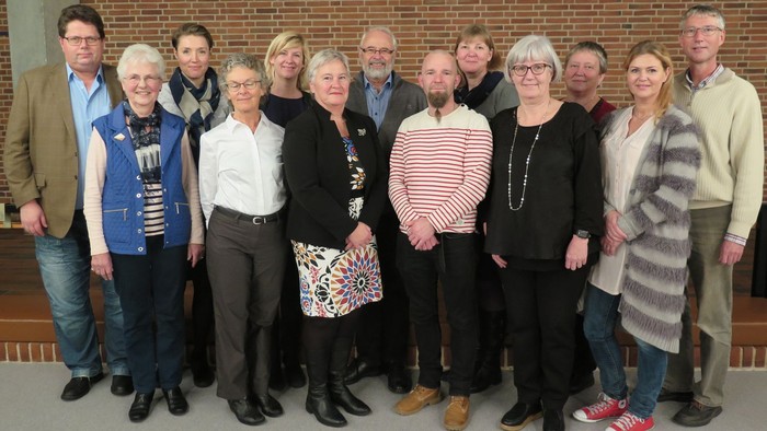 En gruppe af 14 personer står sammen til et fællesskud i et rum med mur af røde mursten.