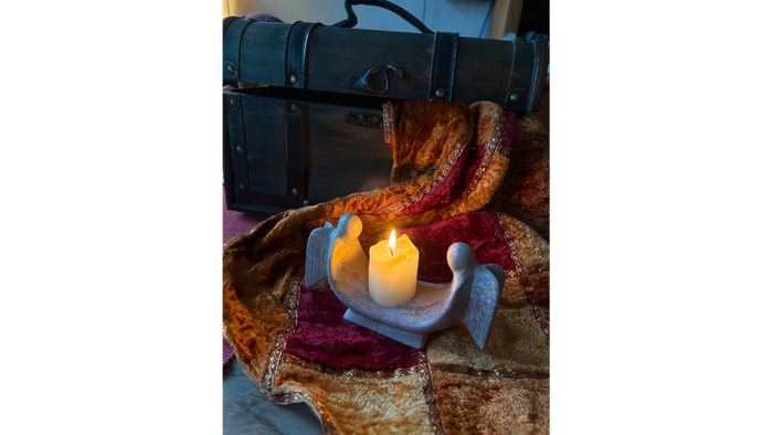 Auf einer Decke, die aus einem Koffer hängt steht ein Kerzenhalter bei dem zwei Engelsfiguren recht und links der Kerze stehen.