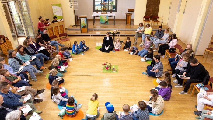 Eine Gruppe von Kindern und Erwachsenen sitzt im Kreis auf dem Boden in einem Raum.