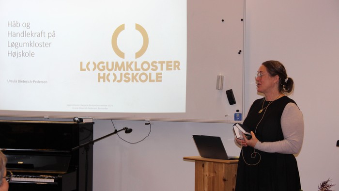 Der ses en storskærm med Løgumkloster Højskoles logo, og ved siden af står forstanderen med en Højskolesangbog i hånden.