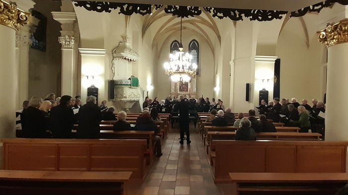 In der Kirche versammeln sich viele Menschen in den Holzbänken, ein großer Kronleuchter hängt von der Decke.