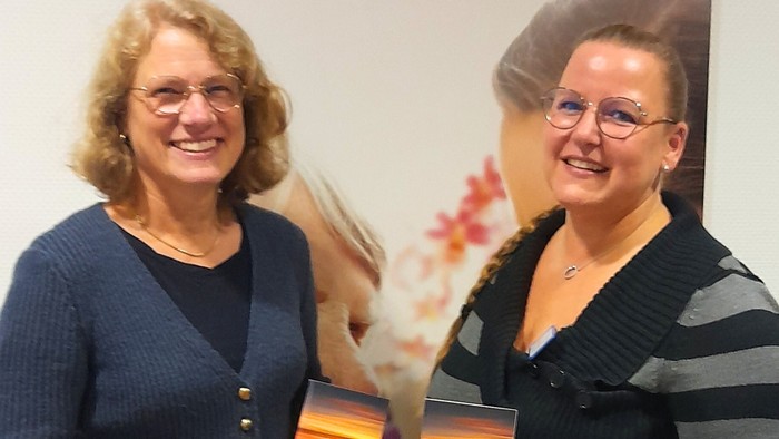 Zwei Frauen stehen vor einem großformatigen Foto und halten jeweils einen Flyer in der Hand.
