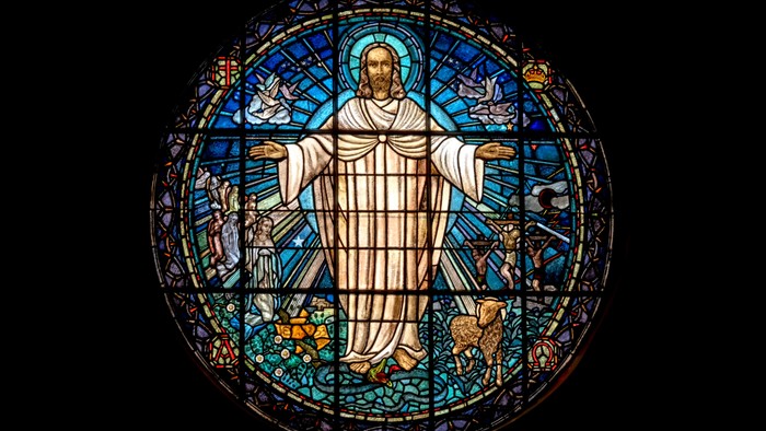 Stained glass window depicting Jesus with outstretched arms, surrounded by colorful designs and symbols.