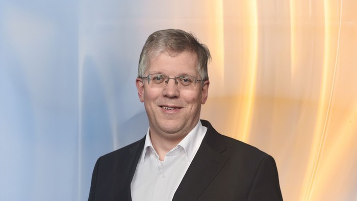 Ein Mann in Anzug und Krawatte lächelt vor einem abstrakt gestreiften Hintergrund.