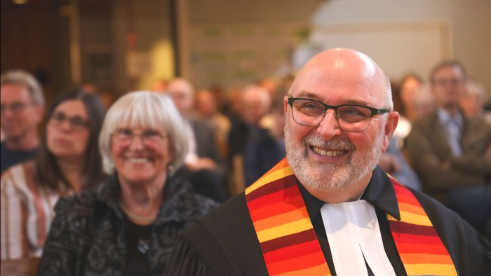 Pfarrer Uwe Stintmann im Talar, sitzend vor der Gottesdienstgemeinde