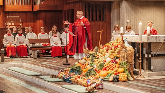 Ein Priester in roter Kleidung segnet eine große Auswahl an Früchten und Gemüse, während eine Gruppe von Menschen zusieht.