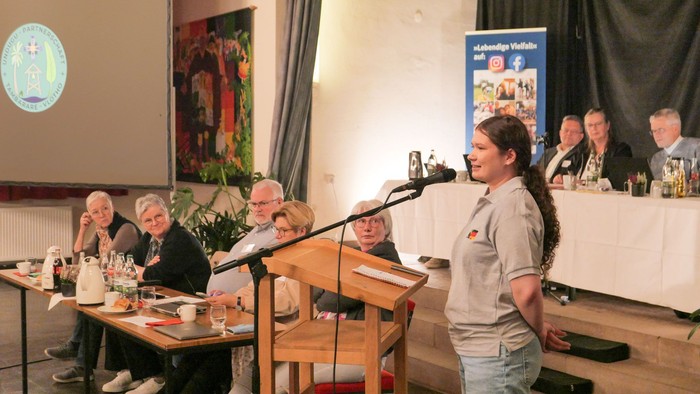 Eine junge Frau spricht am Mikrofon vor zwei Tischreihen mit mehreren Personen. Im Hintergrund eine Leinwand und ein Plakat des Kirchenkreises Vlotho.
