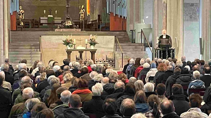 Große Menschenmenge in einer Kirche, viele sitzen, einige stehen, Pfarrer predigt am Altar