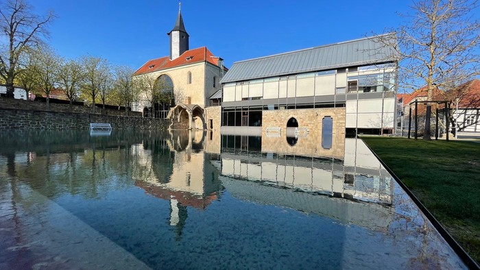 Ein modernes Gebäude mit einem großen Uhrwerk neben einem ruhigen Gewässer und einem Kirchturm im Hintergrund