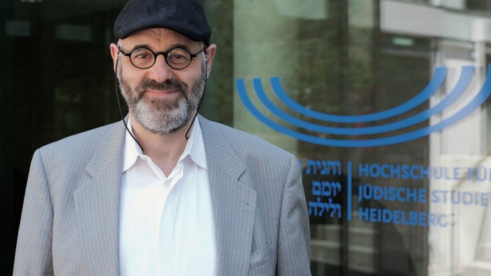 Ein Mann mit Bart, runder Brille und schwarzer Mütze vor einem Fenster mit Aufschrift