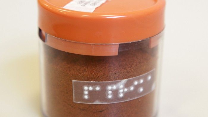 Wer seine Gewürze – hier Paprika – mit Blindenschrift kennzeichnet, kann sie leicht unterscheiden. Foto einer kleinen, transparenten, runden Kunststoffdose mit orangenem Deckel, die ein rotbraunes Pulver enthält. Auf der Dose klebt ein transparentes Etik