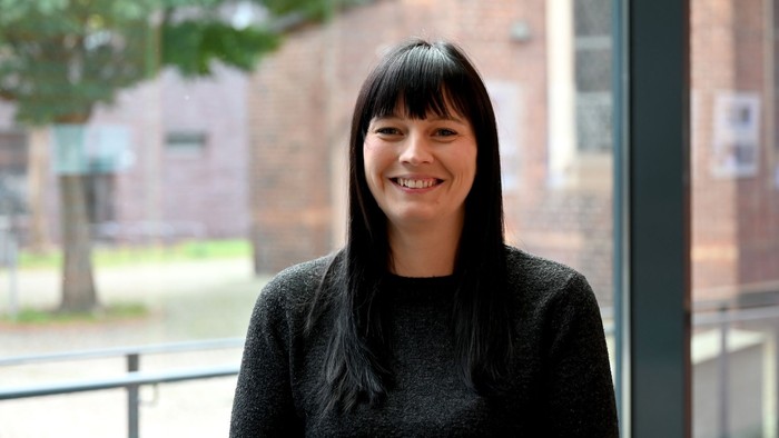 Eine junge Frau mit dunklem Haar lächelt vor einem Backsteingebäude.