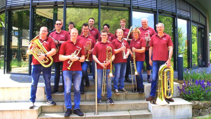 Ein Posaunenchor in roten Polohemden posiert mit Instrumenten auf einer Treppe vor einer Glasfassade