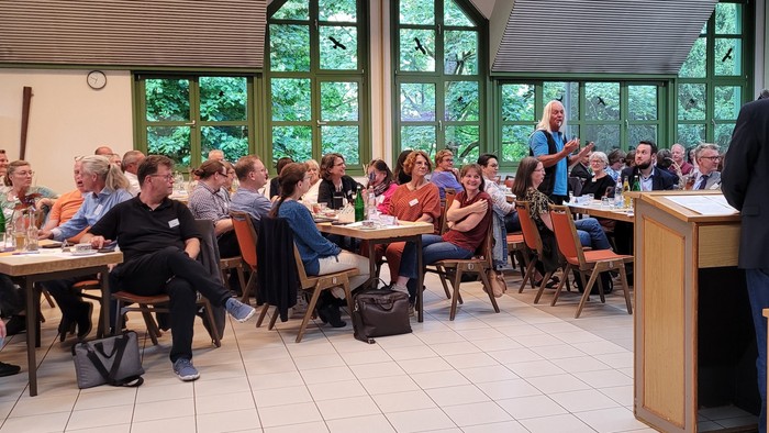 Man sieht viele Menschen an Tschen sitzen. Sie lauschen aufmerksam einem stehenden Mann mit weißen langen Haaren. Im Vordergrund ein Mann mit schwarzem Jacket am Rednerpult.