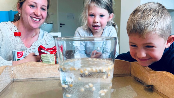 Melanie Hoyer (links im Bild) ist Erzieherin und beobachtet mit zwei Kindern (ein Mädchen und ein Junge) aus ihrem Forscher*innen-Team die tanzenden Maiskörner im Wasserglas. Alle drei lächeln. Das Experiment ist geglückt.
