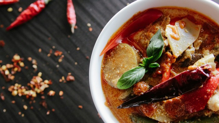 Eine Nahaufnahme von einem roten Thai-Curry in einer weißen Schüssel, garniert mit frischen Basilikumblättern und getrockneten Chilis auf der Oberfläche. Daneben liegen verstreute getrocknete rote Chilis und Chiliflocken auf einem dunklen Holztisch.