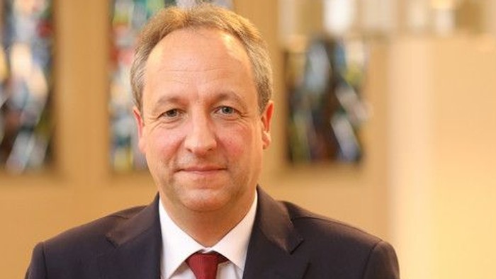 Ein Mann im Anzug mit Krawatte posiert vor einem unscharfen Hintergrund.