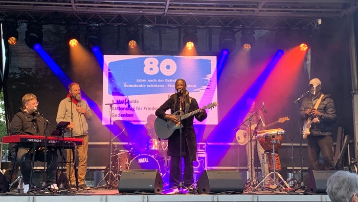 Eine Band spielt auf einer Bühne mit bunten Lichtern und einem großen Bildschirm im Hintergrund.