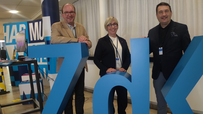 Drei Personen posieren hinter großen blauen Buchstaben.