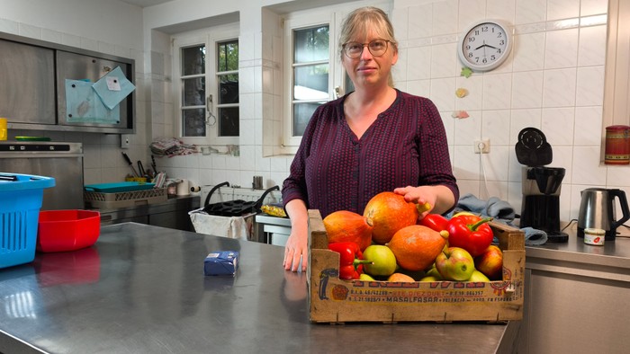 Person steht vor einer Holzkiste voller verschiedener Früchte wie Äpfel, Birnen und Paprika.
