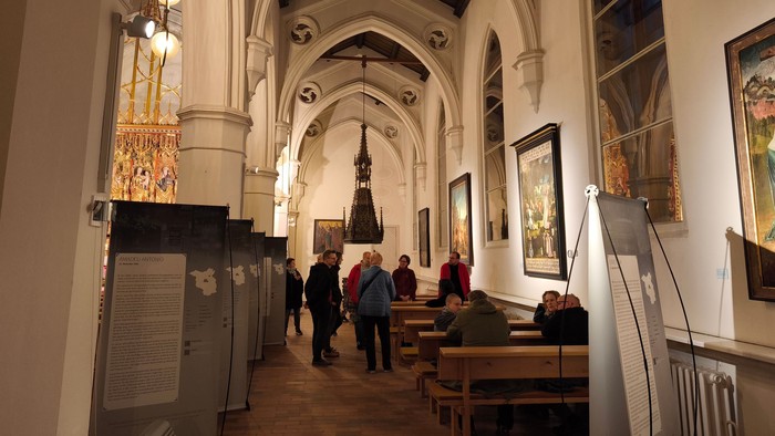 Kircheninnenraum mit hohen Gewölben und Besuchern, die auf Holzbänken sitzen oder stehen.