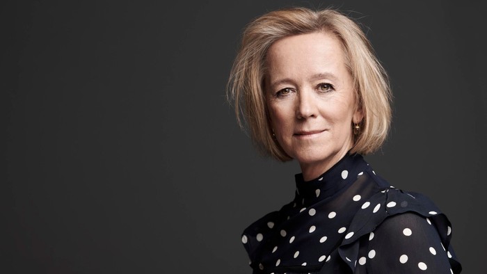 En kvinde med kort, blond hår og polkadot-højtælt sitter i en mørk, udfaset baggrund.