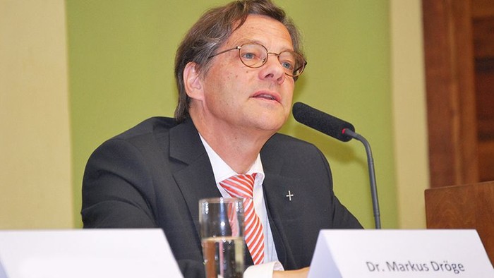 Mann in Anzug spricht hinter Mikrofon an Tisch.