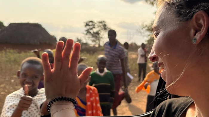 Eine europäische junge Frau winkt aus einem Bus einigen afrikanischen Kindern und Erwachsenen zu. Diese lächeln zurück. Im Hintergrund eine Savannenlandschaft.
