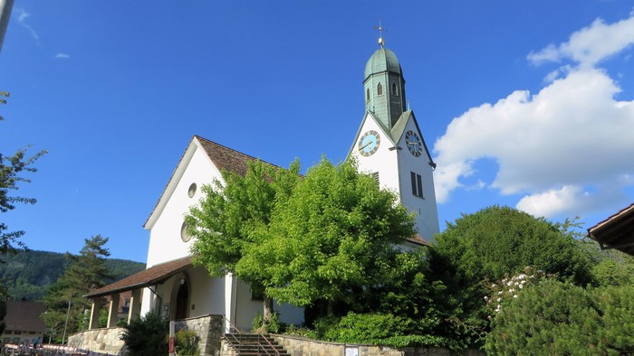 Weiße Kirche mit grünem Turm und Uhr, umgeben von Bäumen und blauem Himmel