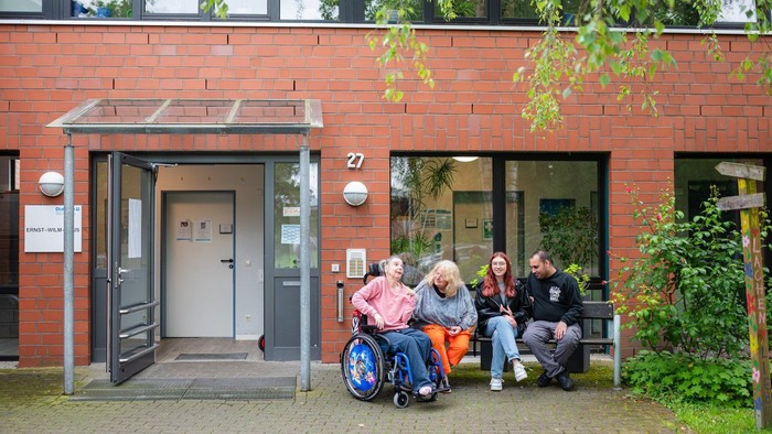 Eine Gruppe von Menschen sitzt vor einem roten Gebäude, eine Person im Rollstuhl.