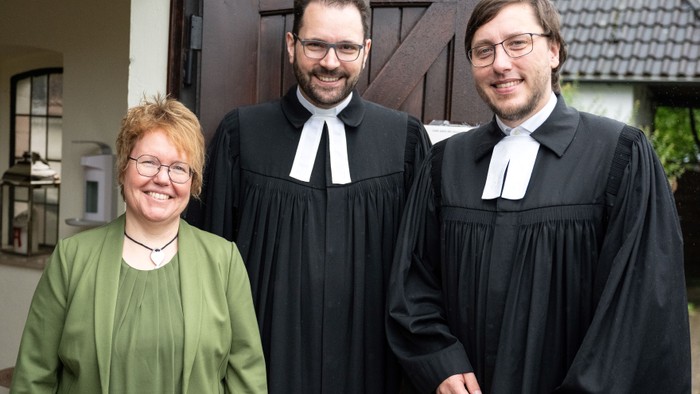 Drei Personen in formeller Kleidung stehen vor einem Haus