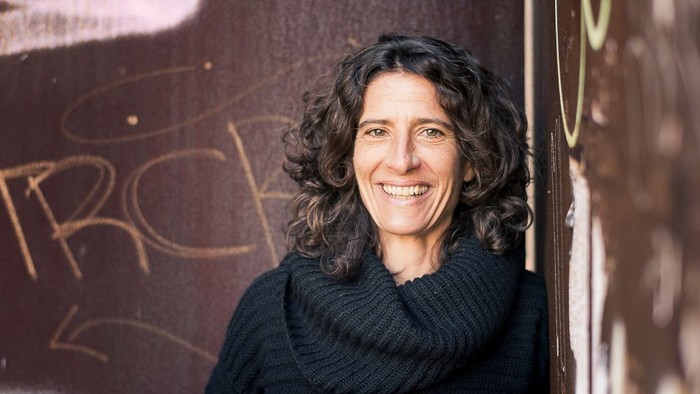 Die Referentin Christiane Groß, mit lockigem Haar, lächelt in die Kamera vor einer Wand mit Graffiti.