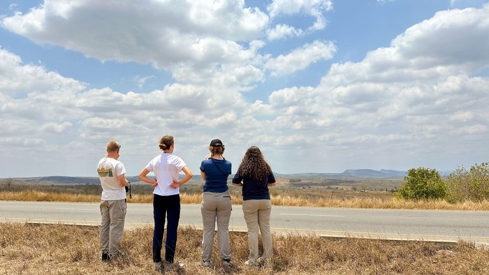Vier Mitglieder der Jugenddelegation stehen am Straßenrand in einer Steppenlandschaft und schauen, von hinten gesehen, in die Ferne. Im Hintergrund Steppe und Berge am Horizont.