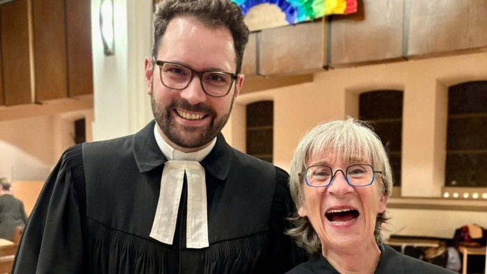 Zwei Personen in schwarzen Roben und weißen Kragen lächeln, eine hält eine Auszeichnung.