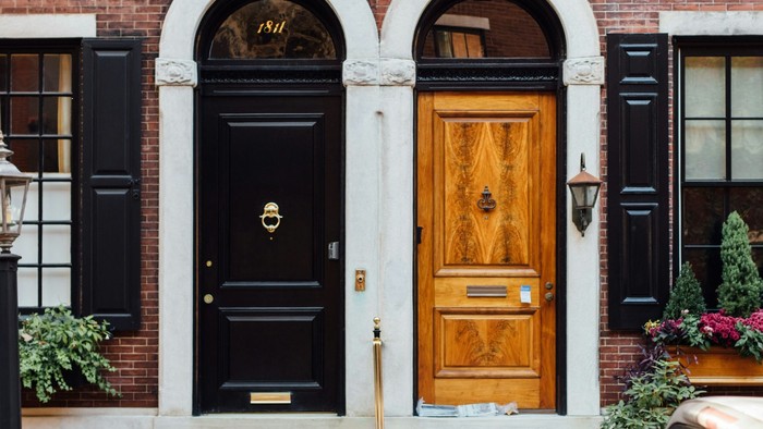 Zwei klassische Haustüren, die linke schwarz lackiert, die rechte klar lasiert, an einer geteilten Backstein-Häuserfront 