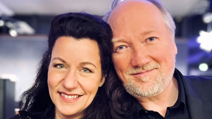 Ein Paar lächelt in die Kamera, die Frau trägt ein schwarzes Kleid, der Mann einen dunklen Anzug