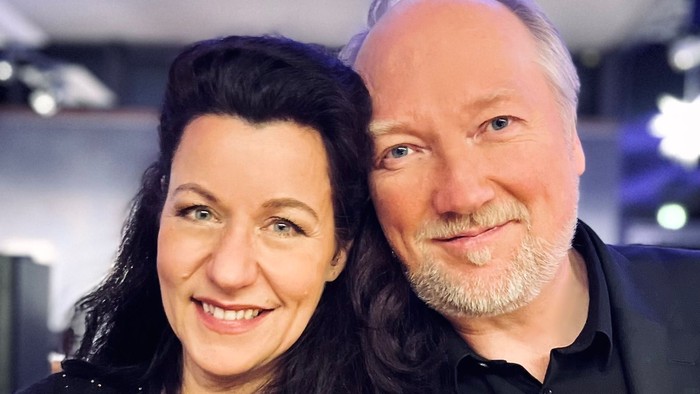 Ein Paar lächelt in die Kamera, die Frau trägt ein schwarzes Kleid, der Mann einen dunklen Anzug