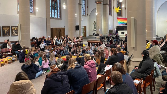 Kircheninneres mit Menschen, die in Reihen sitzen, einige halten Papiere in den Händen, und im Hintergrund ist eine Regenbogenflagge zu sehen.