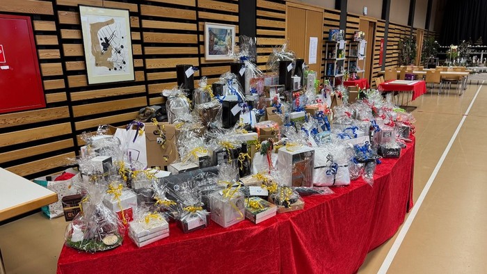 Bord med forskellige genstande på udstilling, heriblandt flasker, æsker og poser, dækket med et rødt bordklæde i et rummeligt lokale.