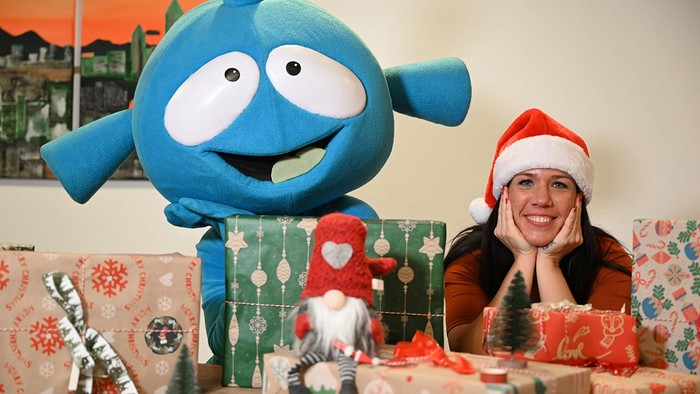 Eine Frau mit einer Weihnachtsmann-Mütze sitzt neben einer großen blauen Figur, umgeben von Weihnachtsgeschenken.