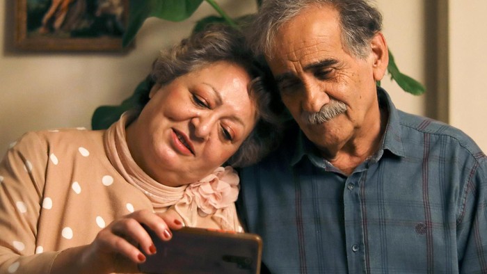 Eine ältere Frau und ein älterer Mann lehnen die Köpfe aneinander und schauen auf ein Handy, das die Frau in der rechten Hand hält. Beide lächeln.