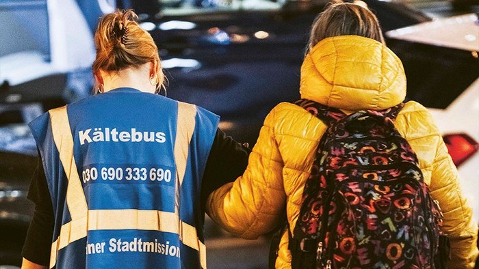 2 Personen von hinten: 1 mit blauer Kältebus-Weste, 1 mit Winterjacke und Rucksack.