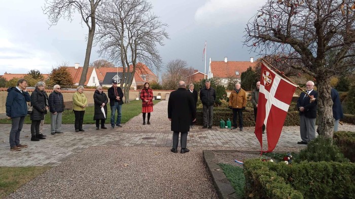 Mennesker ved en mindehøjtidelighed på en kirkegård. Dannebrog og fanebærer er også at se.